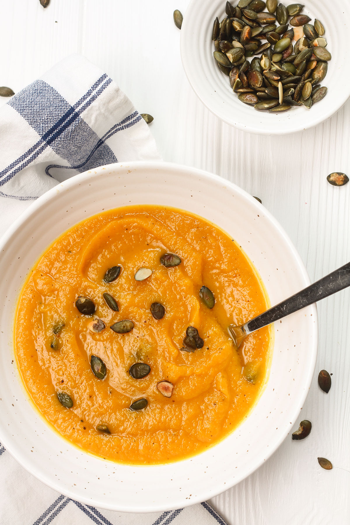 A bowl of pumpkin soup on a white background with a small bowl of pumpkin seeds.