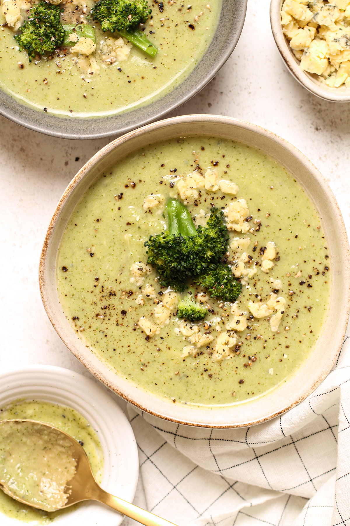 Two bowls of broccoli and stilton soup with a bowl of crumbled stilton for topping.