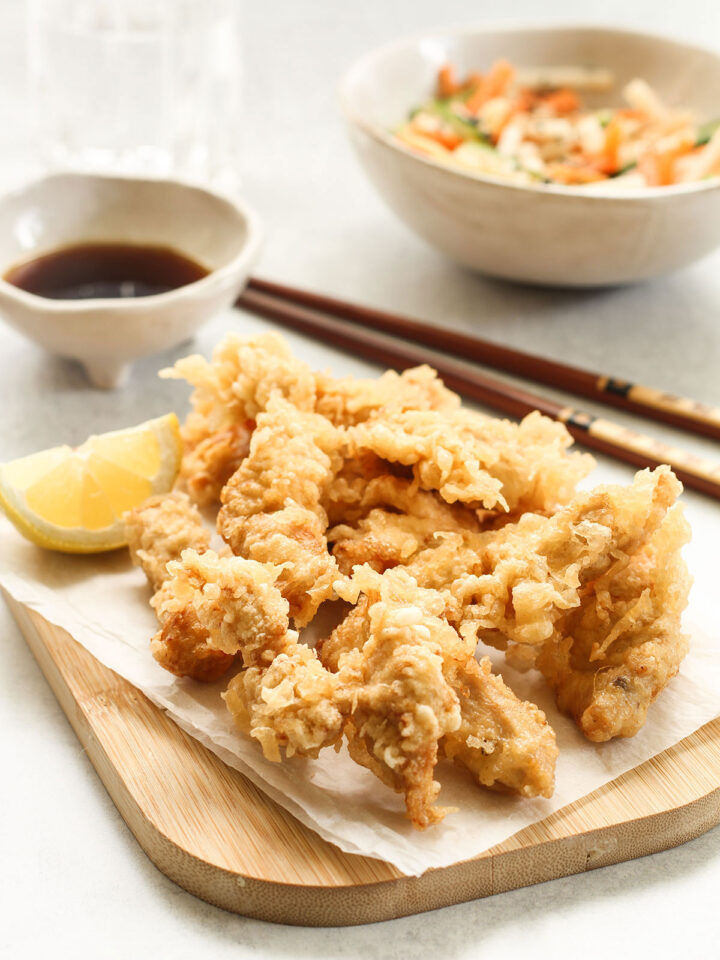 Chicken Tempura with a slice of lemon on a wooden board and a bowl of ponzu dipping sauce, a salad bowl and chopsticks in the background.