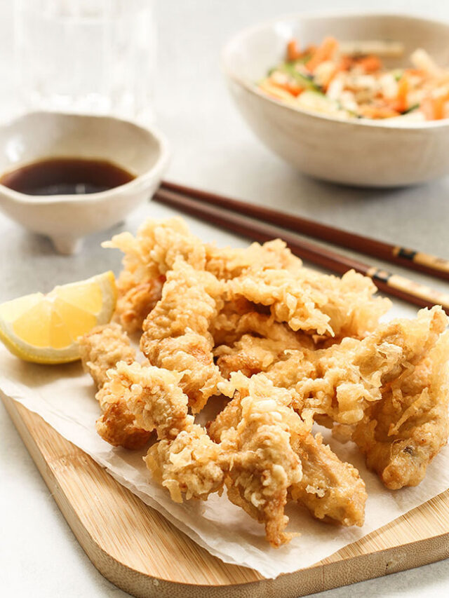 Chicken Tempura with a slice of lemon on a wooden board and a bowl of ponzu dipping sauce, a salad bowl and chopsticks in the background.