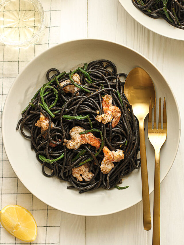 Two white plates of black squid ink pasta topped with samphire and cooked prawns with a small dish of samphire and a slice of lemon on the side.