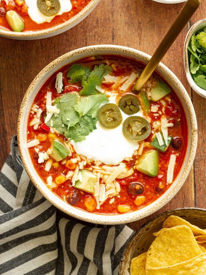 A bowl of Mexican Bean Soup on a wooden table background with Jalapeno, cilantro, cheese and avocado toppings and side dishes of chopped avocado, cilantro and tortilla chips.