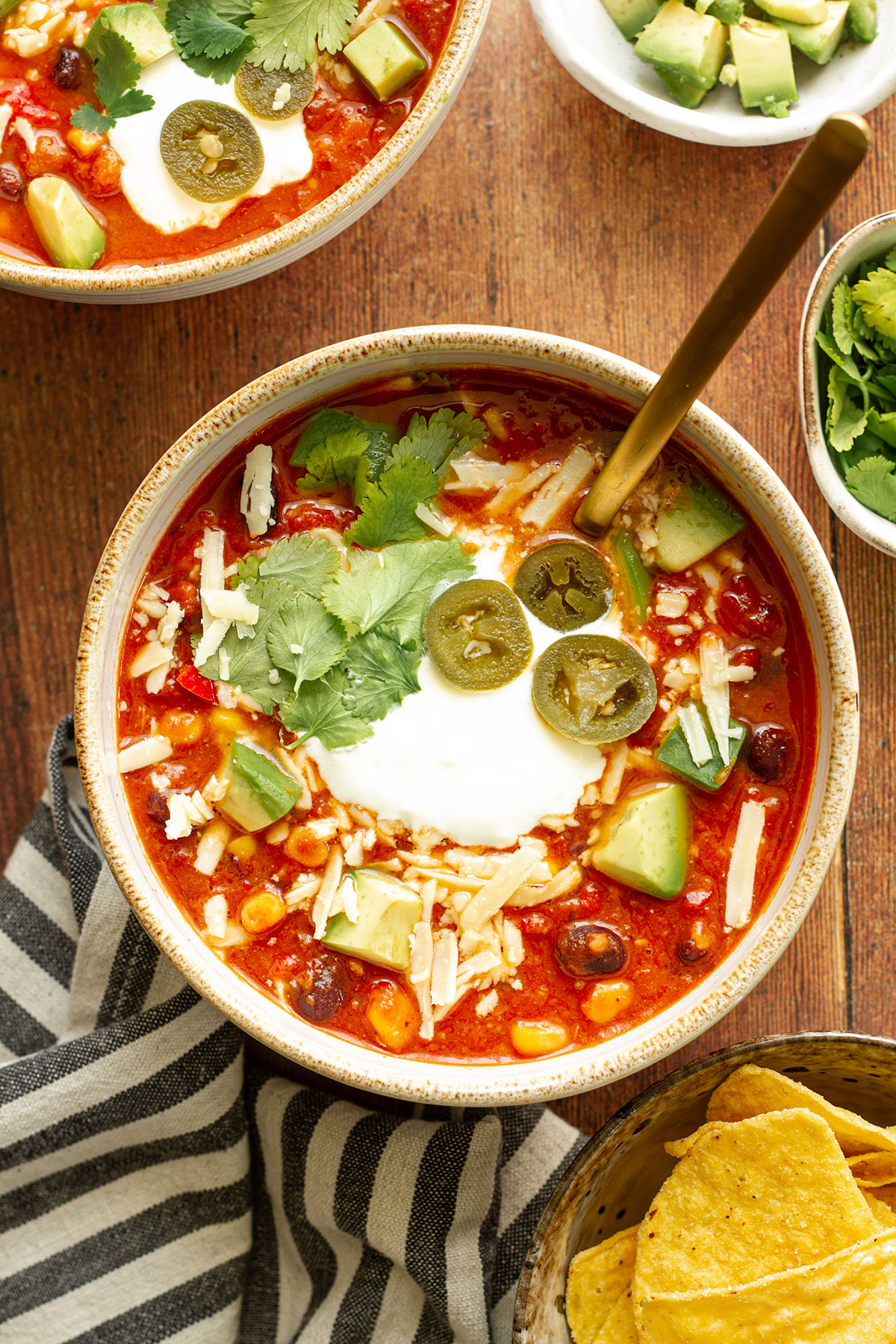 A bowl of Mexican Bean Soup on a wooden table background with Jalapeno, cilantro, cheese and avocado toppings and side dishes of chopped avocado, cilantro and tortilla chips.