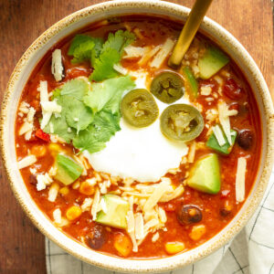 Close up of a bowl of Mexican Bean Soup on a wooden table background, topped with jalapeno, cilantro, cheese and chopped avocado.