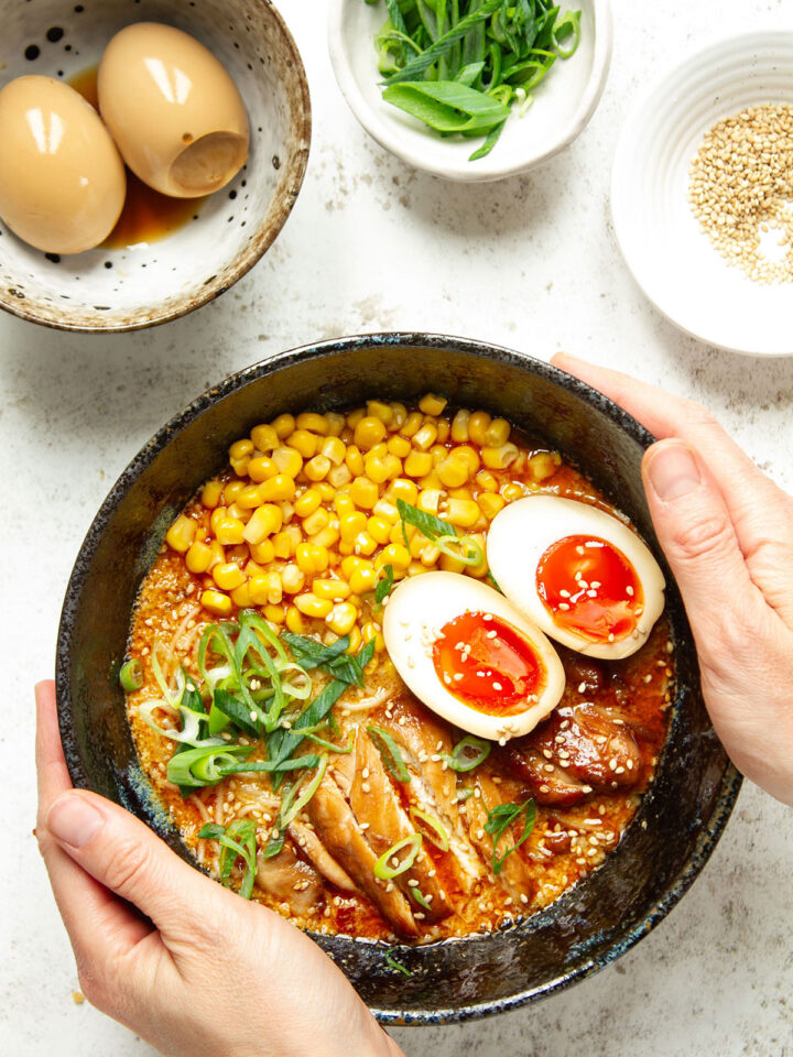 Hands holding a bowl of miso ramen on a white background with three additional bowls containing ramen eggs, sesame seeds and sliced scallions.