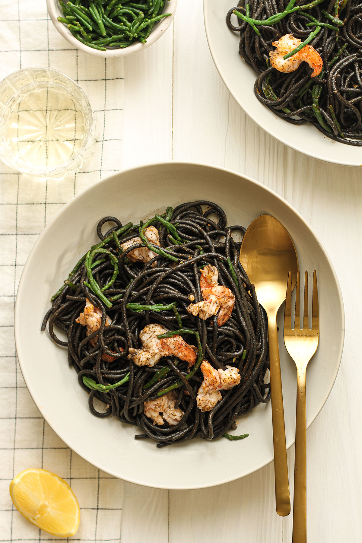 Two white plates of black squid ink pasta topped with samphire and cooked prawns with a small dish of samphire and a slice of lemon.