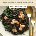 A white plate of black squid ink pasta topped with samphire and cooked prawns with a small dish of samphire and slices of lemon on the side.