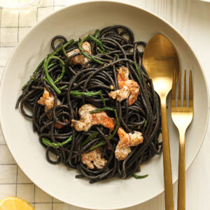A white plate of black squid ink pasta topped with samphire and cooked prawns with a slice of lemon.