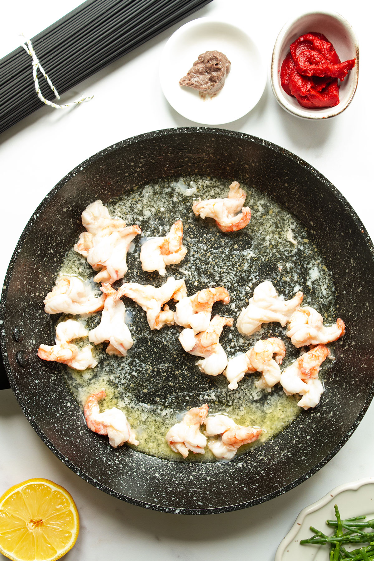 A pan of shrimps cooking for a few minutes until opaque with with small dishes contaning samphire, tomato puree and anchovy paste and a half lemon.