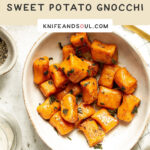 A bowl of sweet potato gnocchi and sage leaves with a spoon on a white table and white check cloth background.