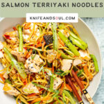 A bowl of Teriyaki Salmon Noodles on a pale blue background with chopsticks and side dish of greens.