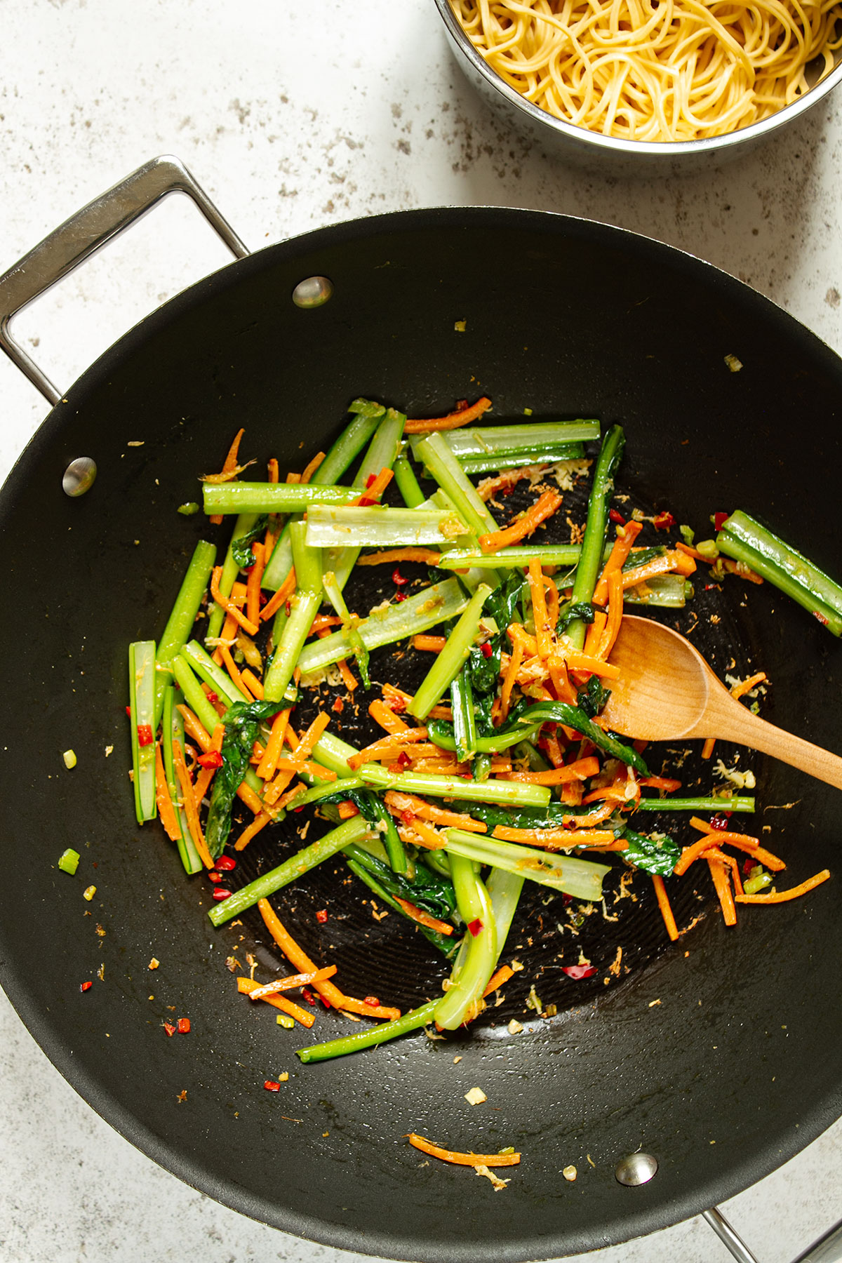 A wok containing cooked vegetables with the noodles on a white background.