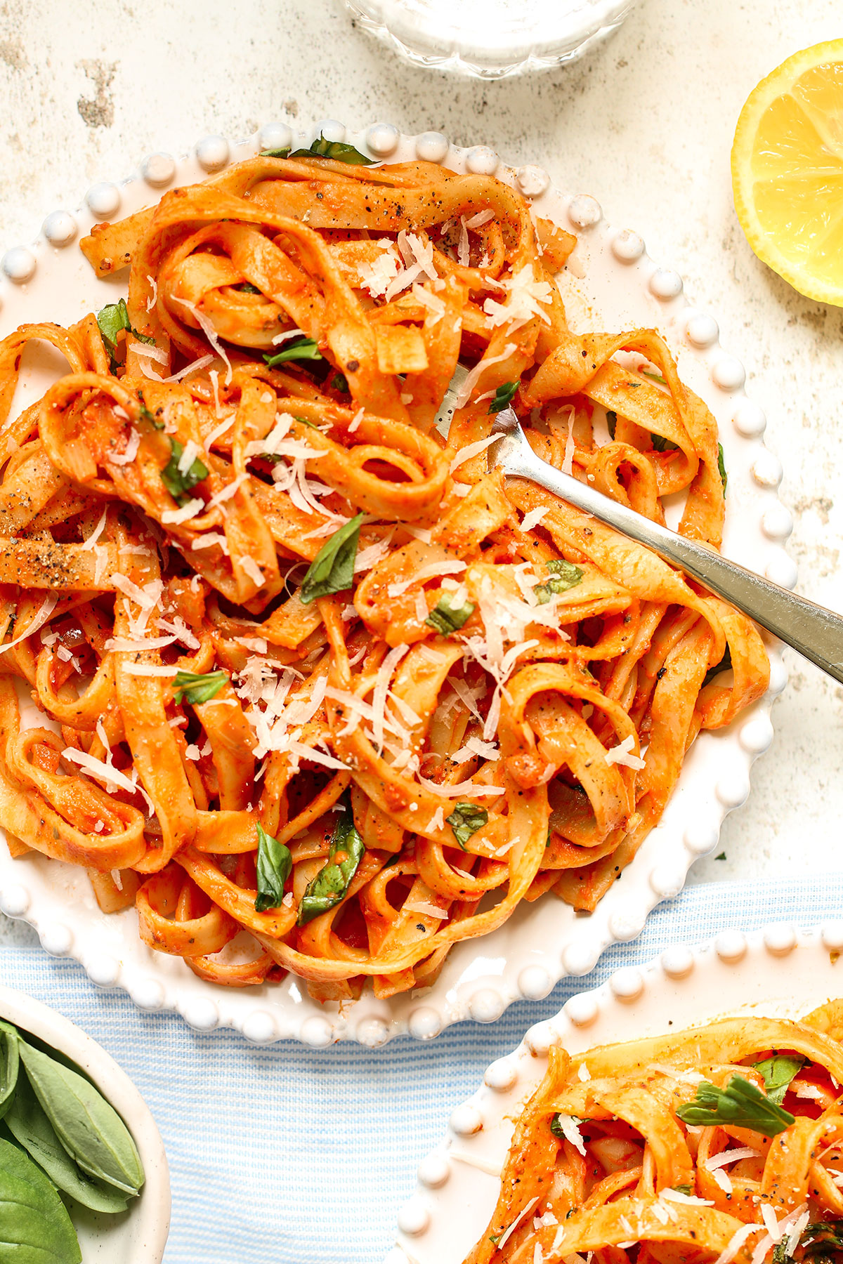 Two bowls of Mascarpone Tomato Pasta on a white background with a fork and sides of basil leaves and half a lemon.