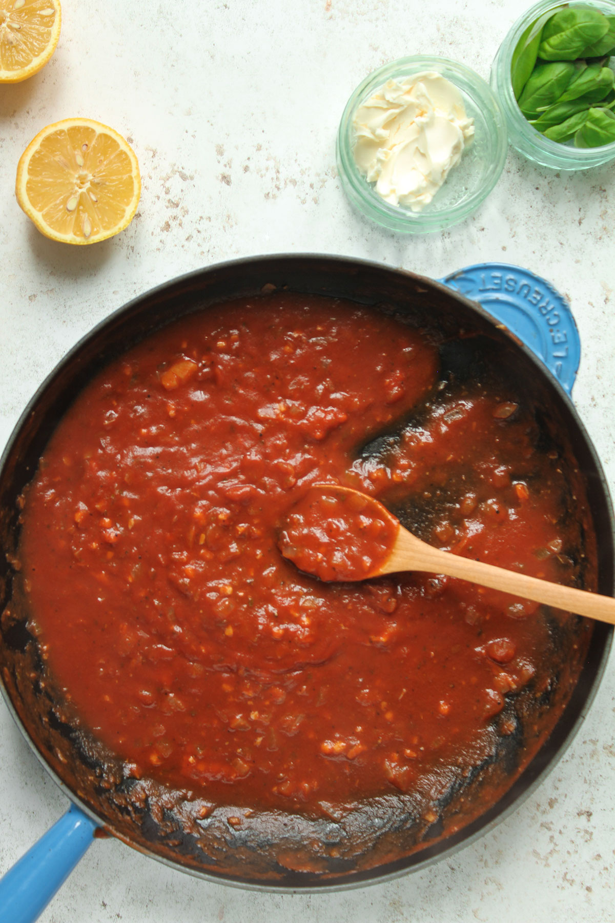 The seasoned passata sauce in a frying pan on a white background and half a lemon, a jar of mascarpone and a jar of basil leaves.