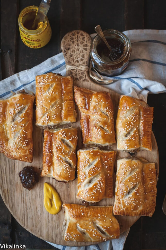 Caramelised onion sausage rolls served on a wooden board with pickle and mustard.