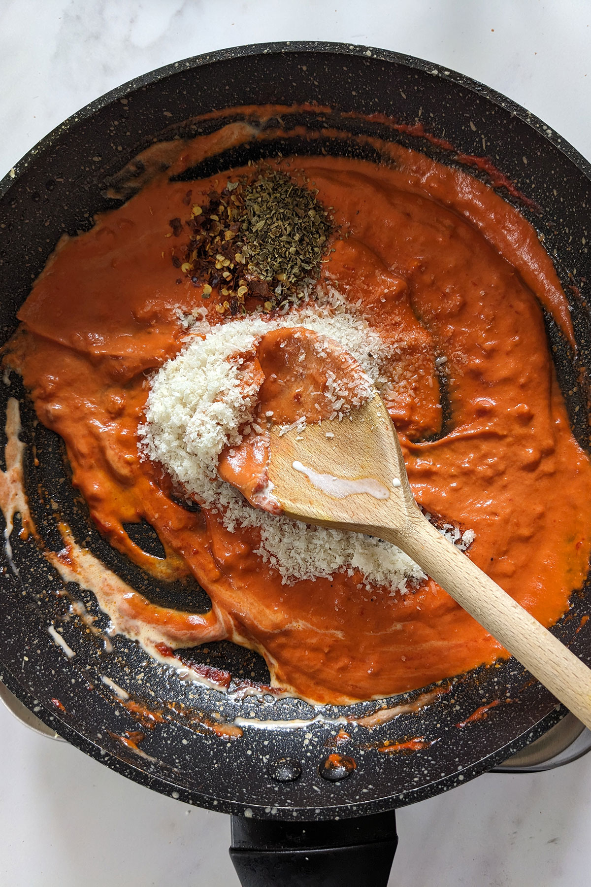 Stirring the Parmesan, red pepper flakes, oregano, salt, and pepper with a wooden spoon in a dark coloured frying pan.