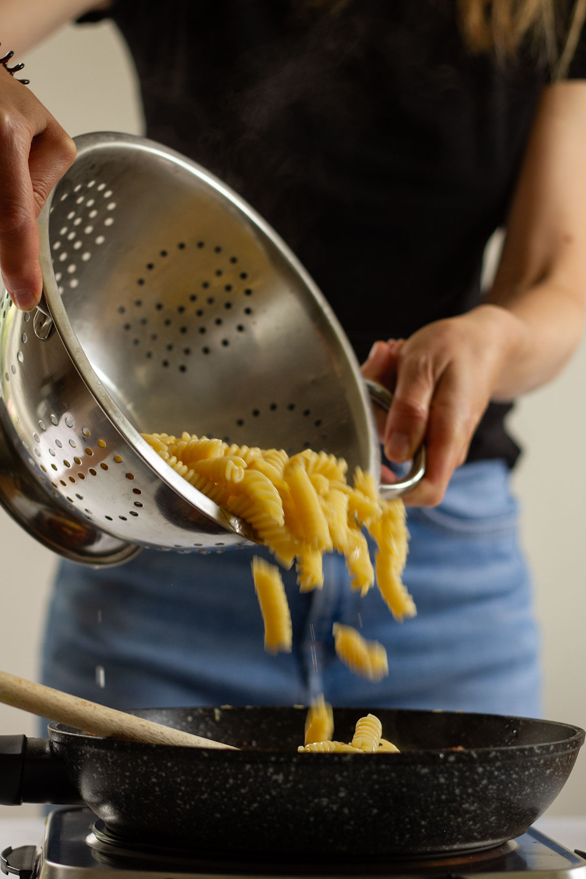 Pouring the cooked pasta from a silver colander into the sauce in a dark coloured frying pan.