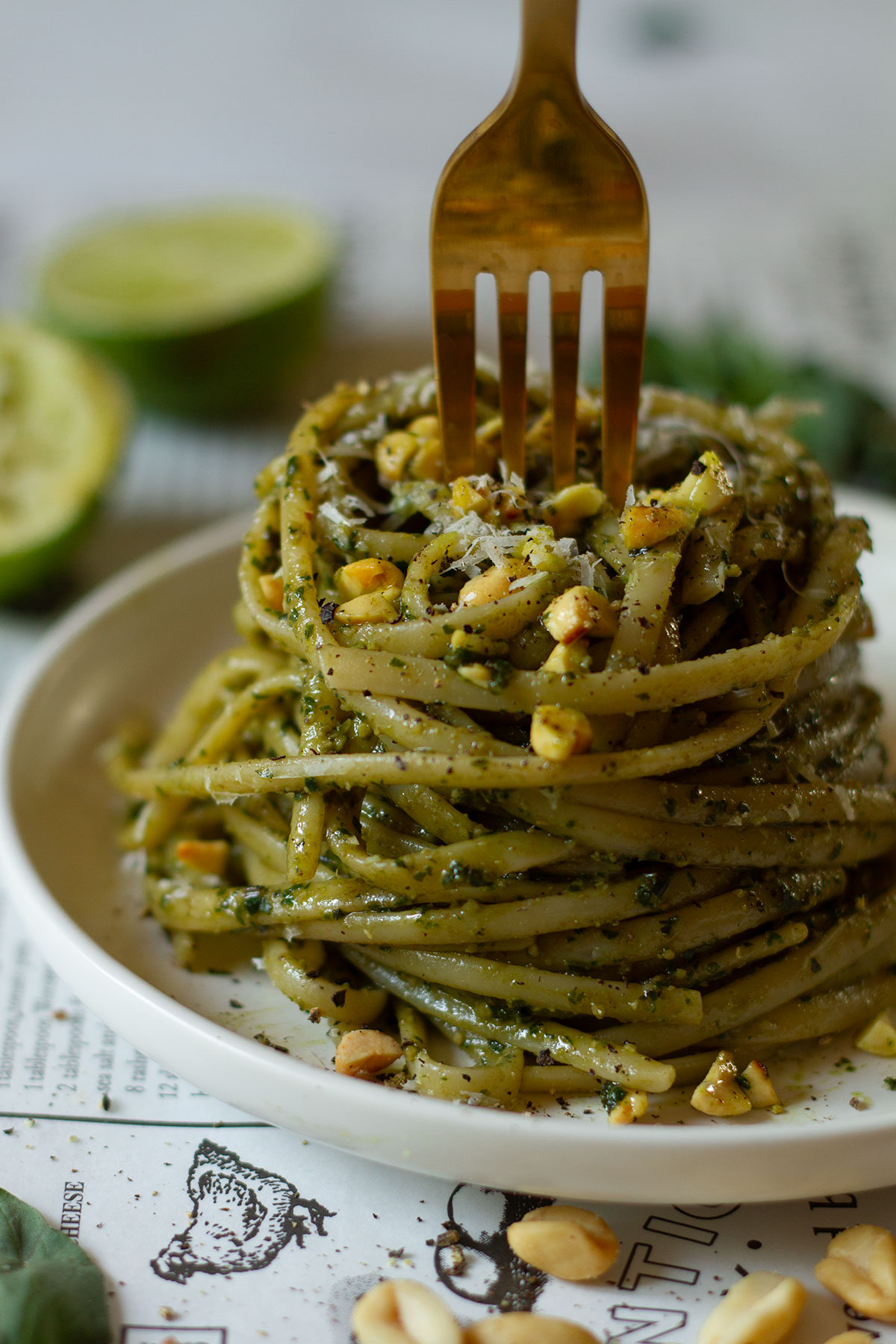 Thai Basil Pesto served with pasta on a white plate on a newspaper themed background with a scattering of peanuts and basil leaf in the foreground and a squeezed lime behind the plate.