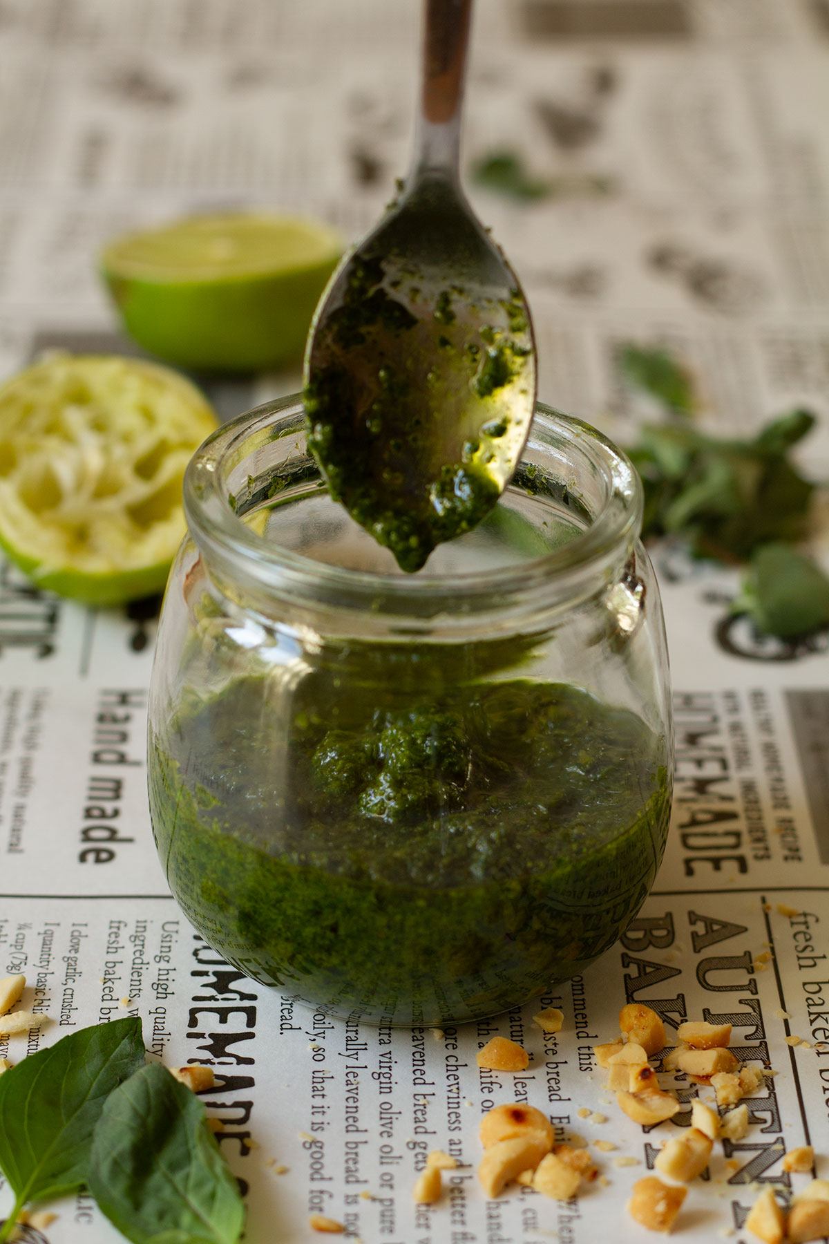 Thai Basil Pesto in a glass jar with a spoon on a newspaper themed background with a scattering of peanuts and basil leaf in the foreground and a squeezed lime behind the jar.