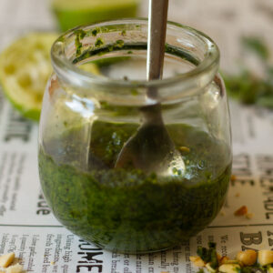 Thai Basil Pesto in a glass jar with a spoon on a newspaper themed background with a scattering of peanuts in the foreground and a squeezed lime behind the jar.