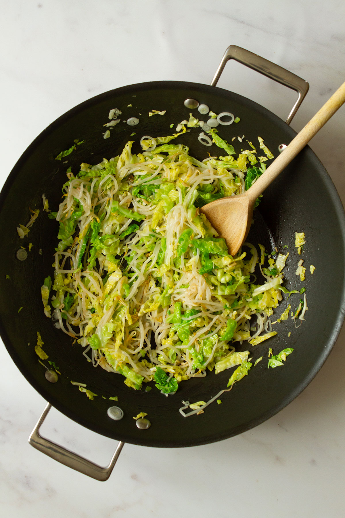 Stirring the fried cabbage, scallions, bean sprouts and garlic in a dark coloured wok using a wooden spoon.
