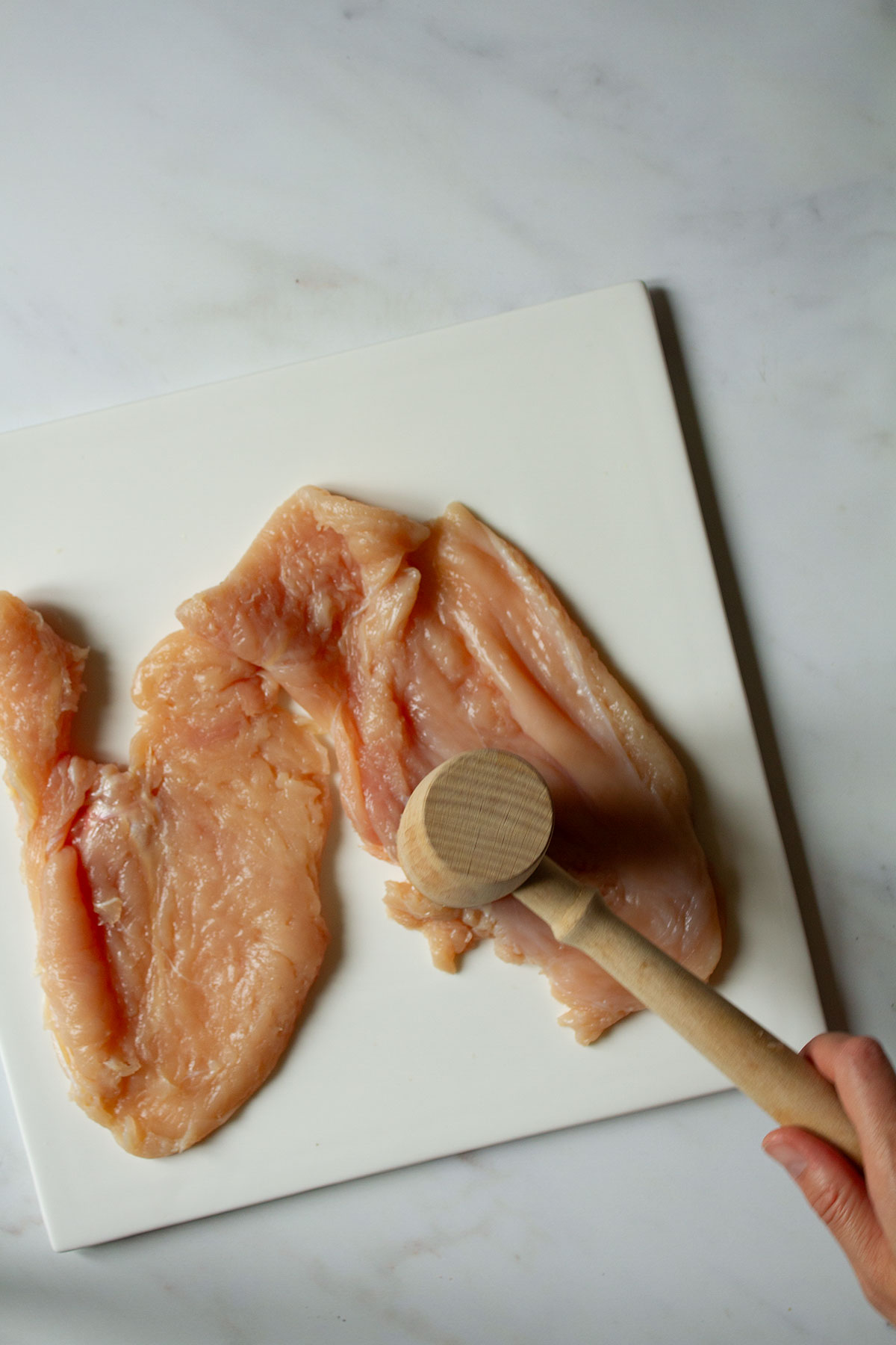 Pounding the chicken breasts with a meat tenderiser on a white surface .