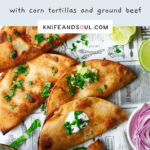 A serving of pan fried tacoes on a newspaper themed background with sides of red onion, guacamole, cilantro and lime wedges.
