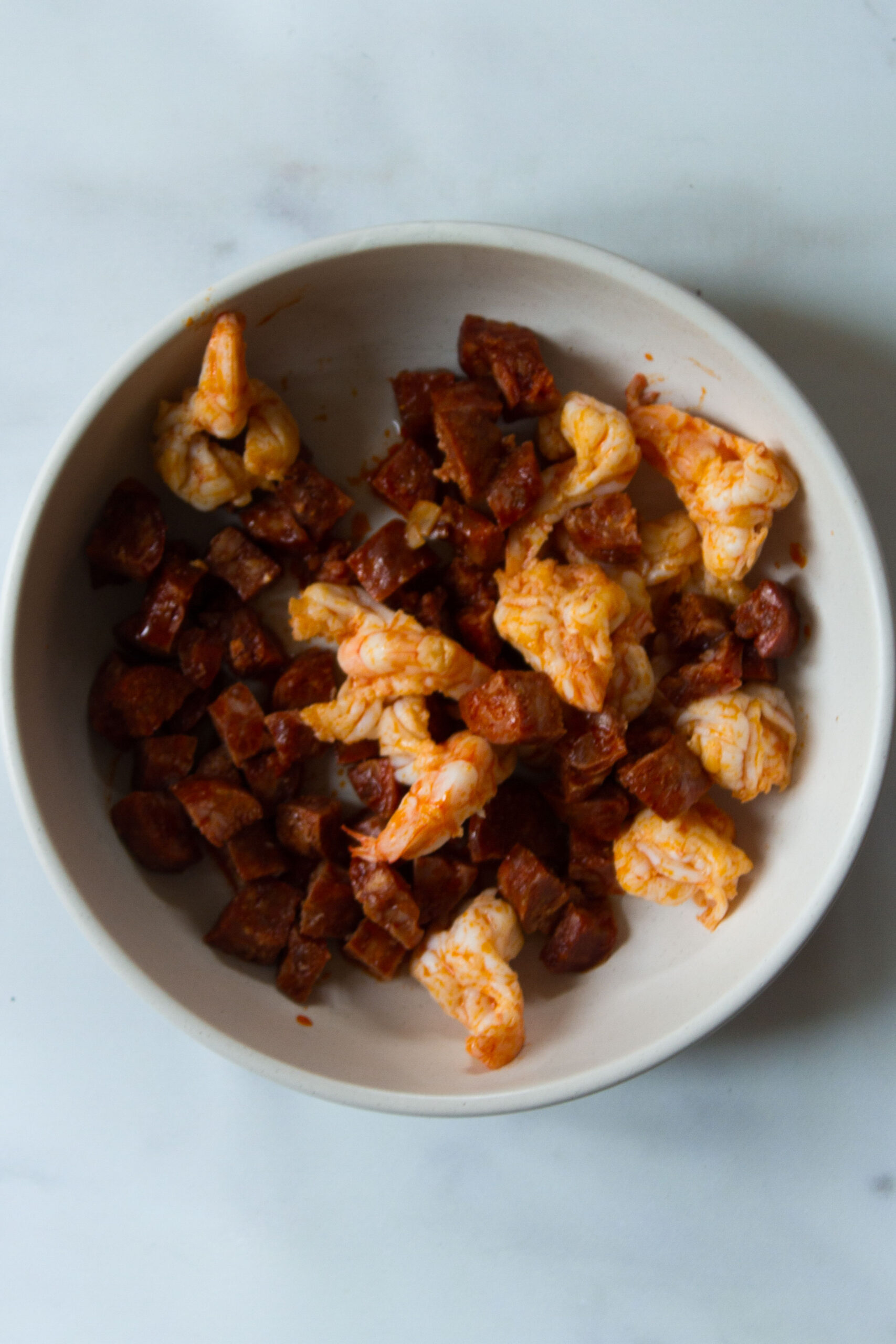 Thr cooked chorizo and prawns set aside in a white bowl on a pale blue background.
