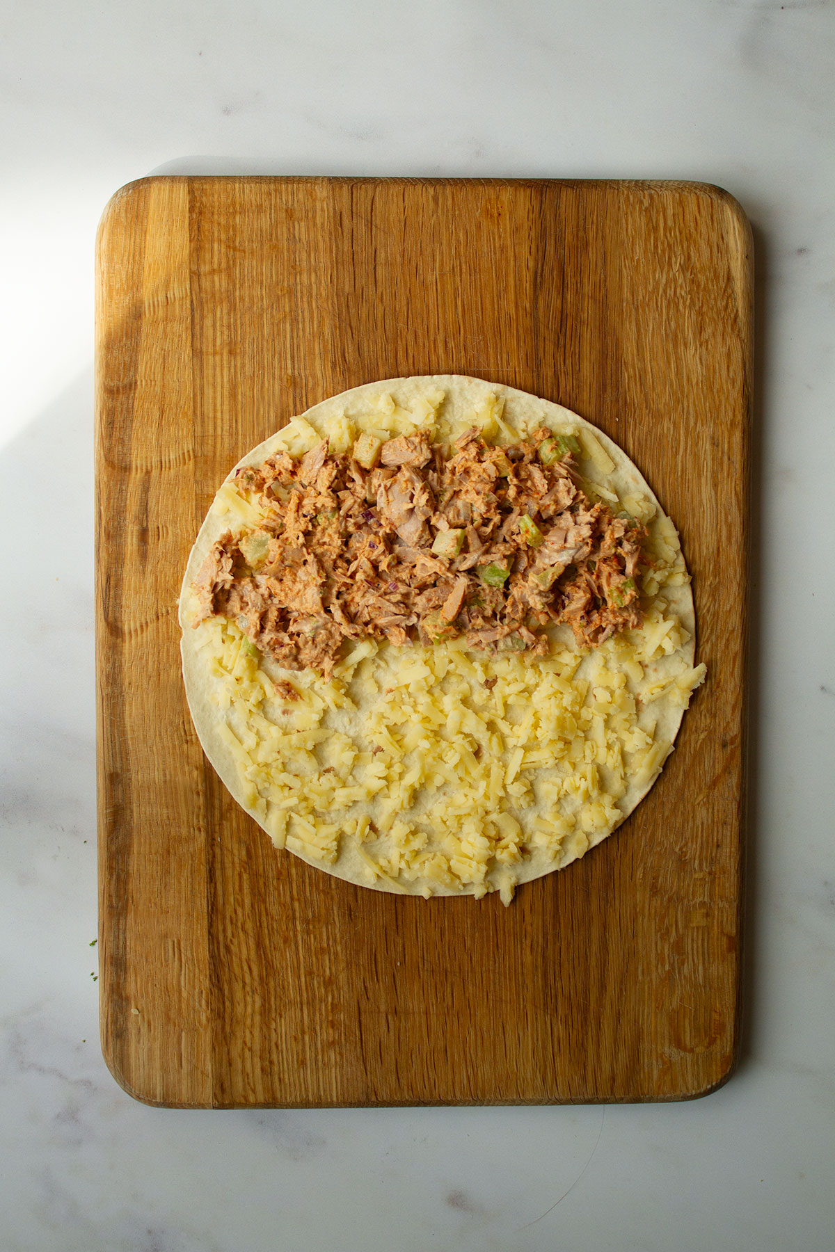 A quesadilla wrap on the wooden board topped with tuna mayo and grated cheese.