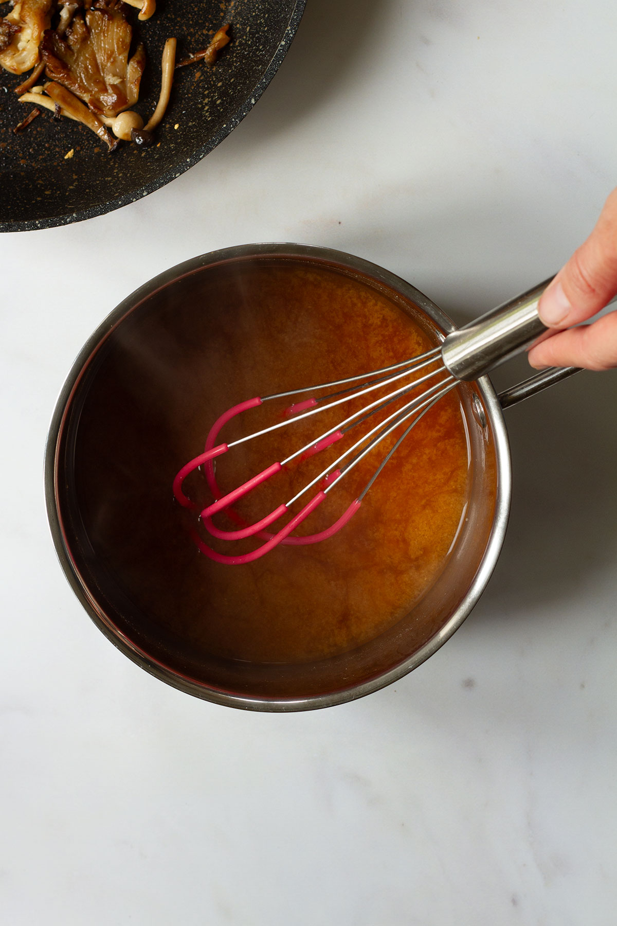 A pan containing miso and dashi with a hand holding a whisk.
