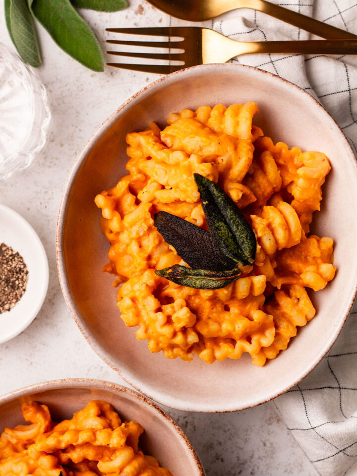 Two bowls of Roast Pumpkin Pasta topped with crispy sage leaves on a white and chequered cloth background.