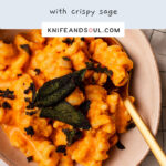 A bowl of Roast Pumpkin Pasta topped with crispy sage leaves on a white and chequered cloth background.