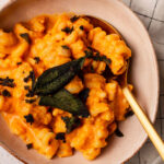 A bowl of Roast Pumpkin Pasta topped with crispy sage leaves on a chequered cloth background.