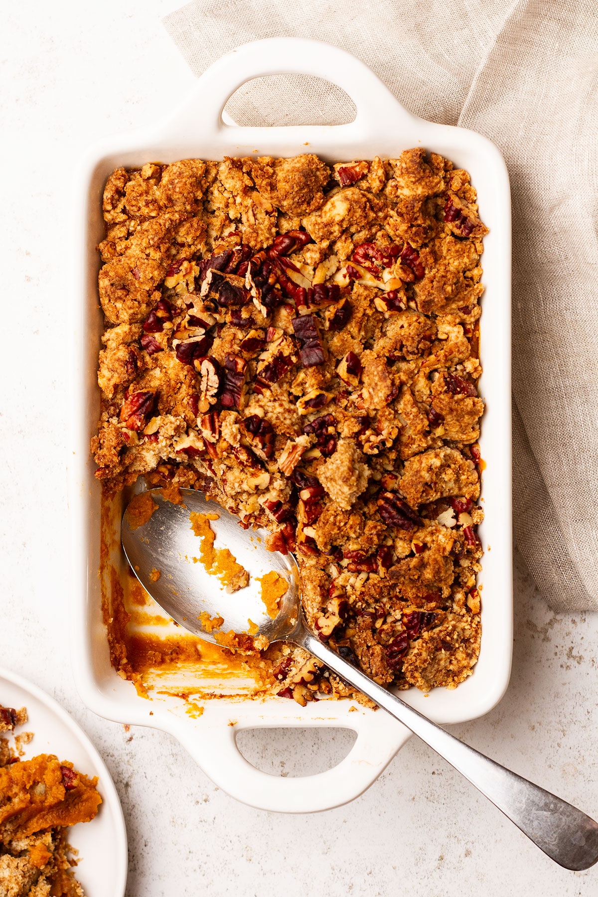 Sweet Potato Casserole being served from a white baking dish with a silver spoon, on a white and linen cloth background.