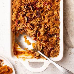 Sweet Potato Casserole being served from a white baking dish with a silver spoon, on a white and linen cloth background.
