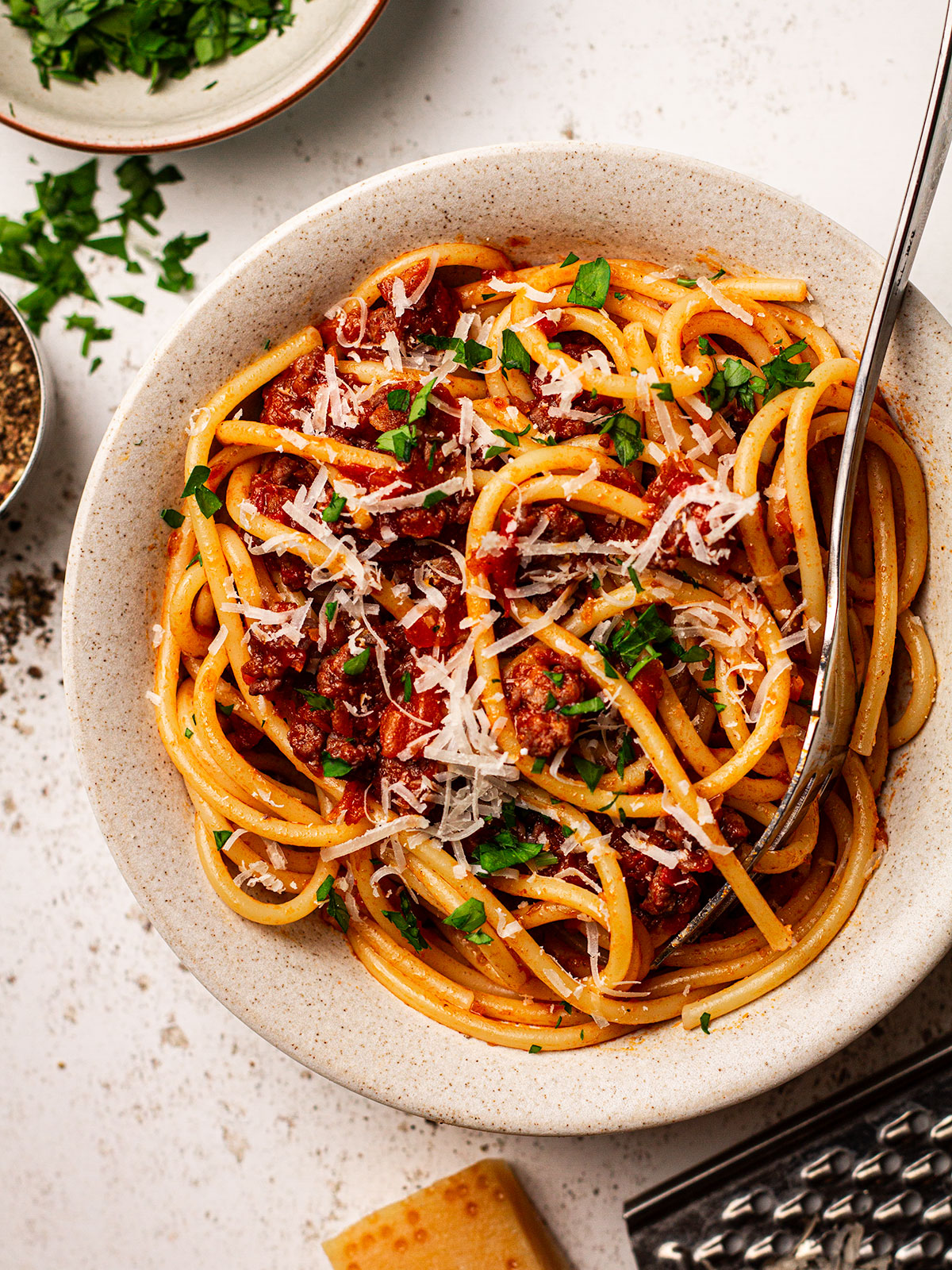 A serving of Spaghetti Meat Sauce (Bolognese) ina white bowl on a white bckgrpound with with freshly grated Parmesan and finely chopped basil on the side.