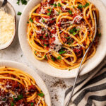 Two servings of Spaghetti Meat Sauce (Bolognese) in white bowls on a white bckgrpound with with freshly grated Parmesan and finely chopped basil on the side.