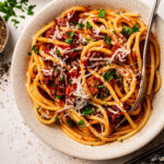 A serving of Spaghetti Meat Sauce (Bolognese) in a white bowl on a white bckgrpound with with freshly grated Parmesan and finely chopped basil on the side.