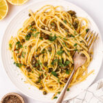 A serving of lemon caper pasta on a white plate with lemons and black pepper on the side.