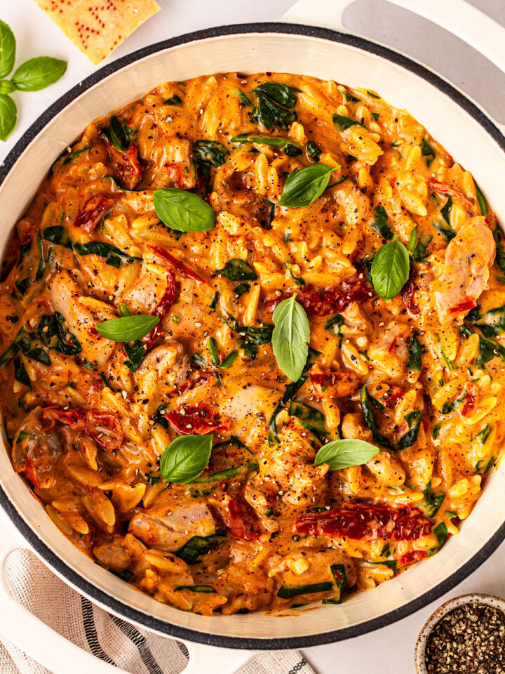 Baked chicken orzo ready to serve with basil leaves on top and pepper and parmesan on the side. In a cream pot on a white background.