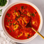 A serving of spicy tortellini soup with chives on the side. In a white bowl on a white background.