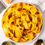A serving of Saffron Pasta in a white bowl on a white background with sides of Parmesan and chopped pistachio.