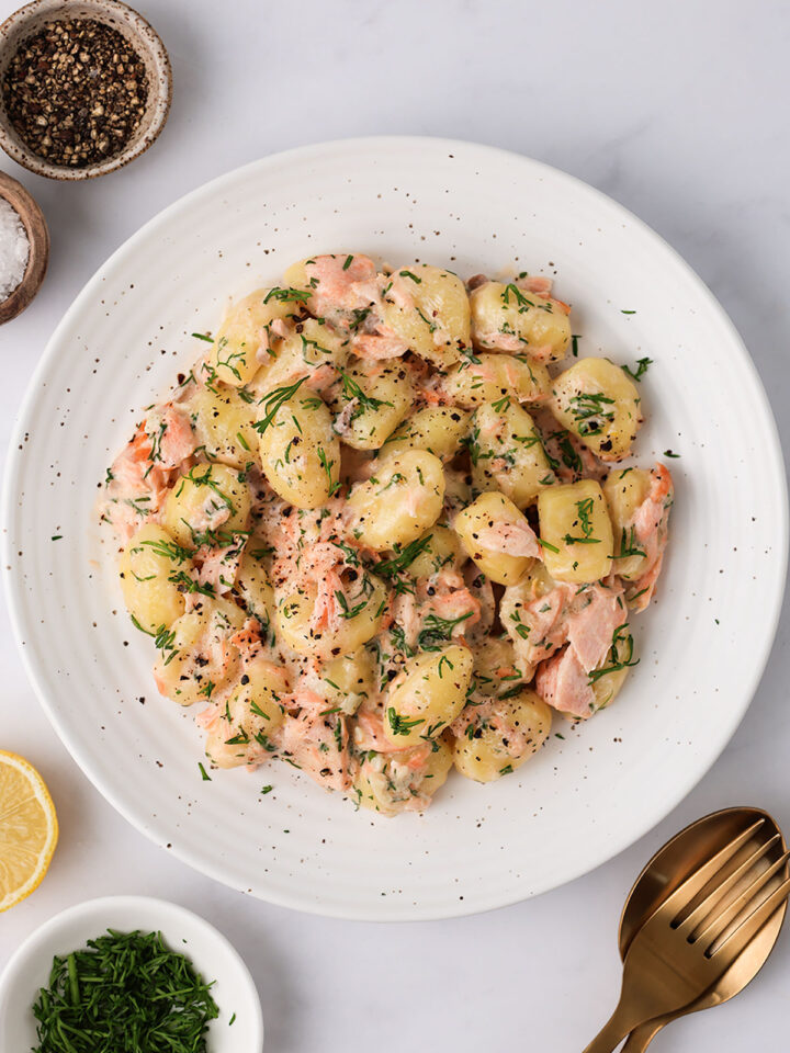 A serving of Creamy Salmon Gnocchi on a white plate with lemon, pepper and dill on the side.