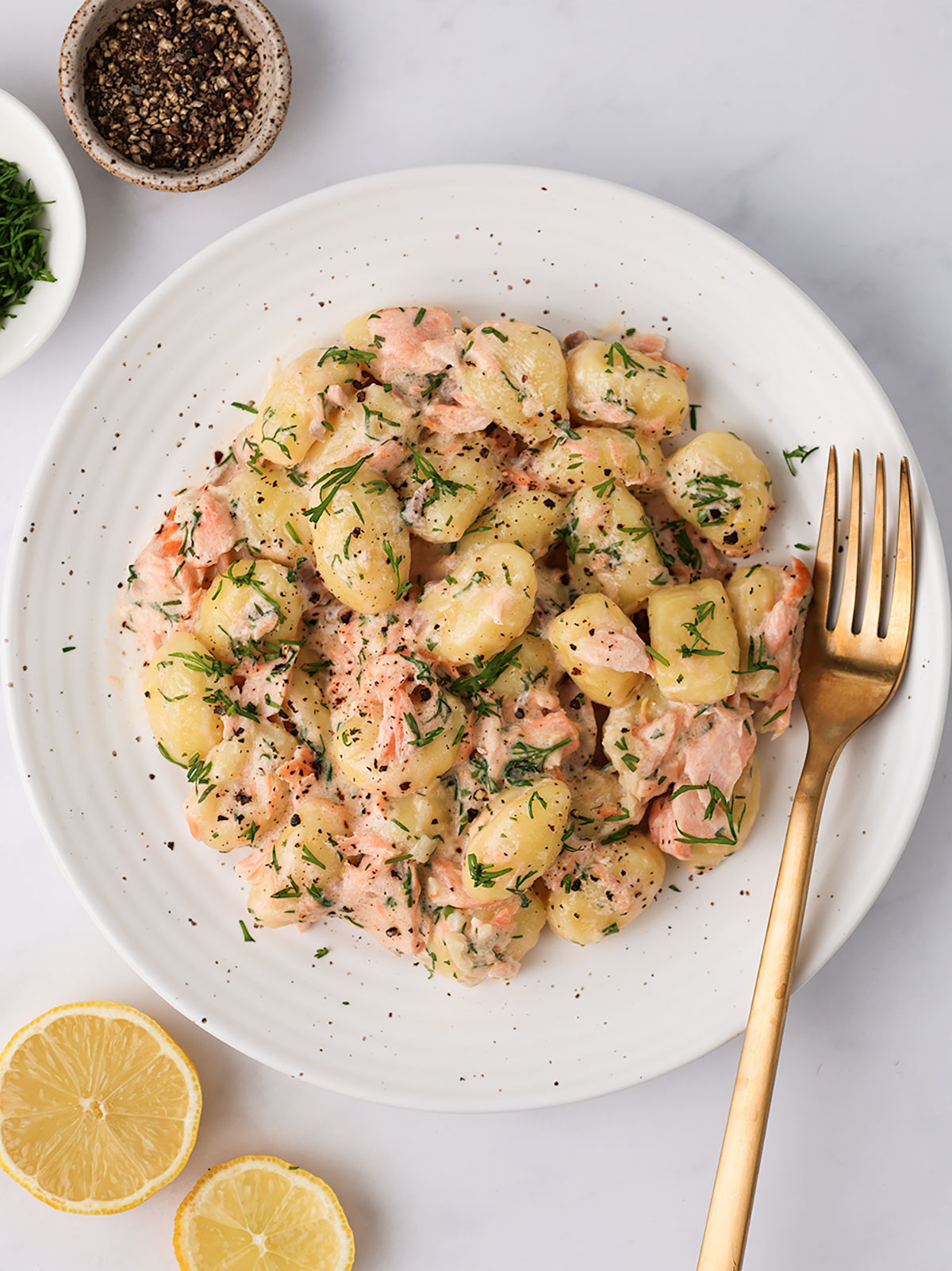 A serving of Creamy Salmon Gnocchi on a white plate with lemon, pepper and dill on the side.
