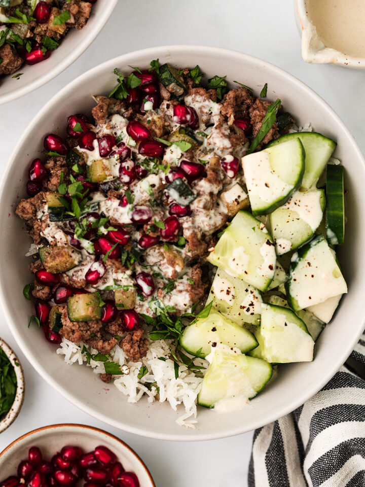 A serving of Ground Lamb Bowl in a white bowl on a white background. With pomegranate seeds and cilantro on the side.