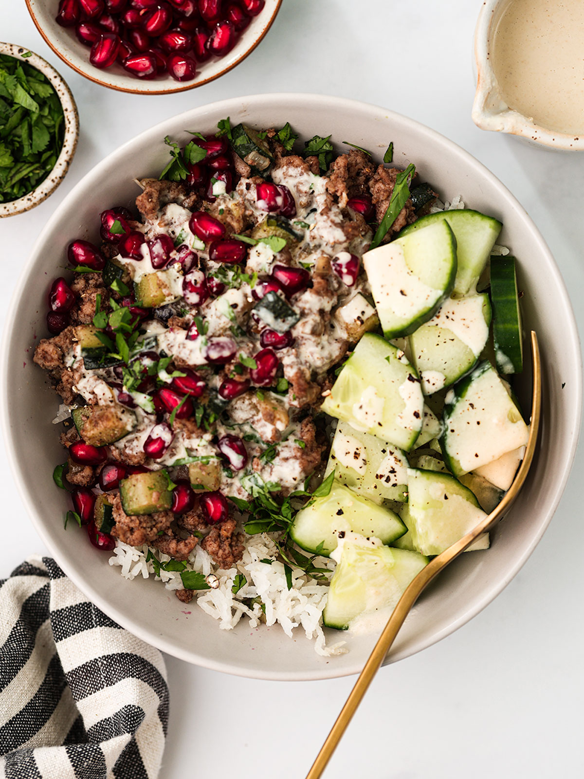 A serving of Ground Lamb Bowl in a white bowl on a white background. With pomegranate seeds and cilantro on the side.