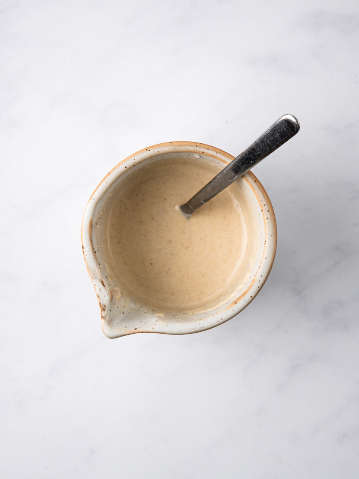 Mixing all tahini sauce ingredients, adding water until you get a drizzleable consistency. In a white bowl on a white background,