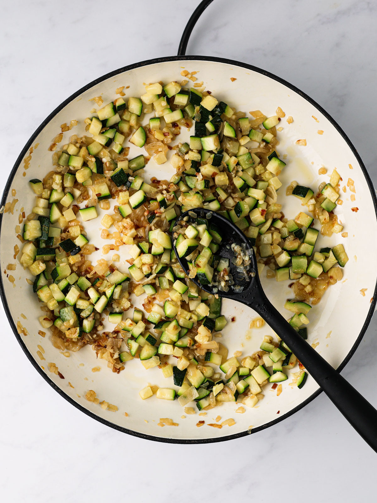 Cooking the zucchini 3-4 minutes until lightly browned. In a white electric skillet with a black spoon on a white background.