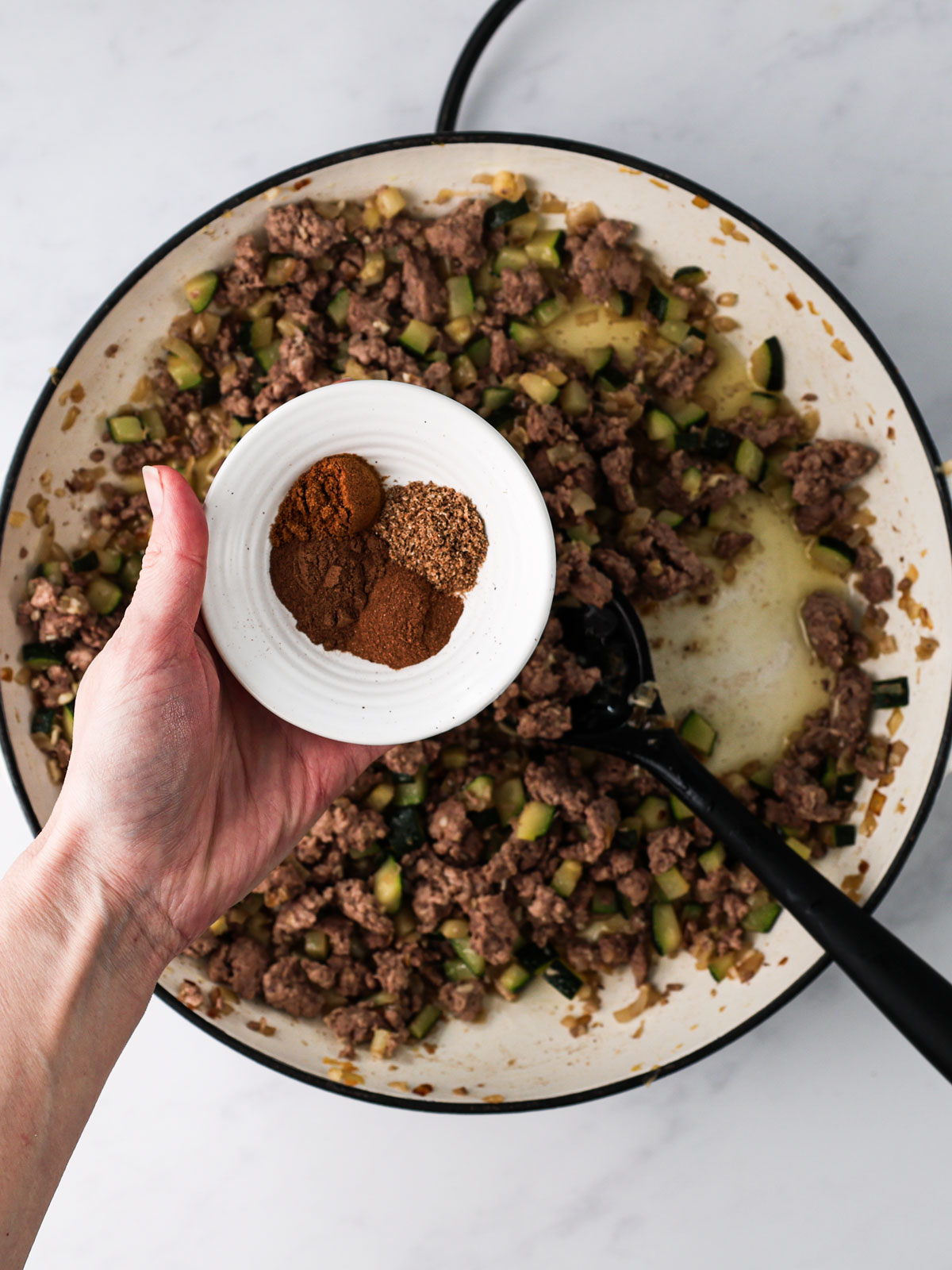 Stir in the ground coriander, allspice, cinnamon, cumin, and salt. Cook 1-2 minutes until fragrant and everything is evenly coated. In a white electric skillet with a black spoon on a white background.
