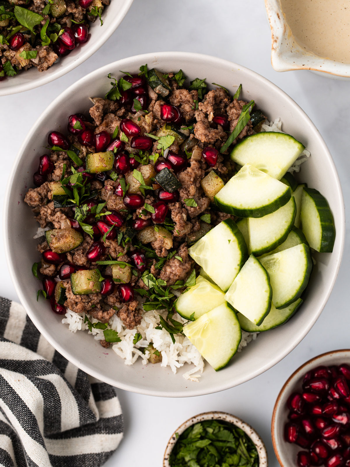 Serve over rice, couscous or orzo. Drizzle with tahini sauce, scatter cilantro and pomegranate seeds on top, and add cucumber on the side. In a white bowl on a white background.